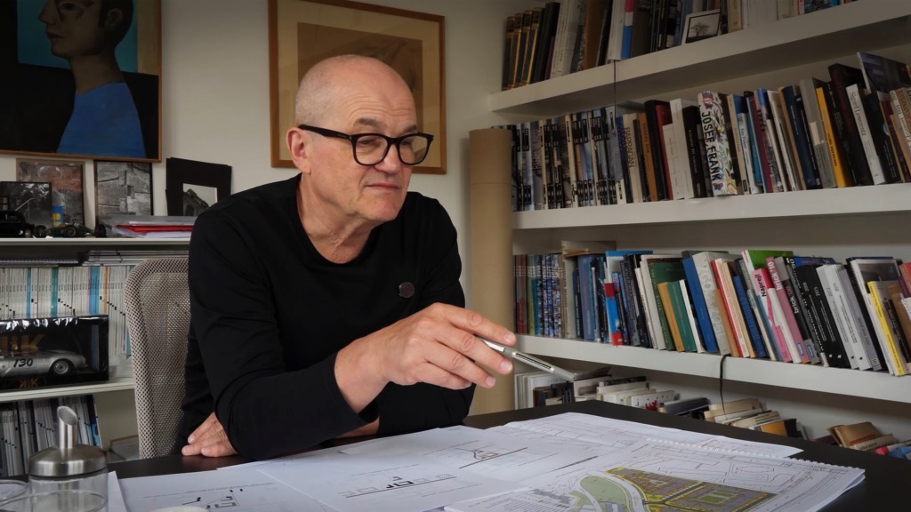 Architekt v brýlích a černém svetru studuje výkresy u stolu s knihovnou v pozadí