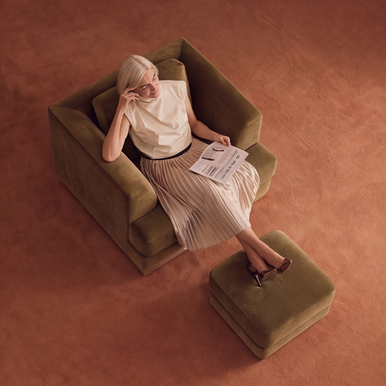 Top view of a woman reading a newspaper in a green armchair on a terracotta rug