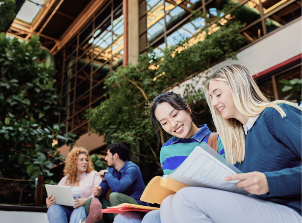 Smiling female students looking through documents in a bright atrium with greenery
