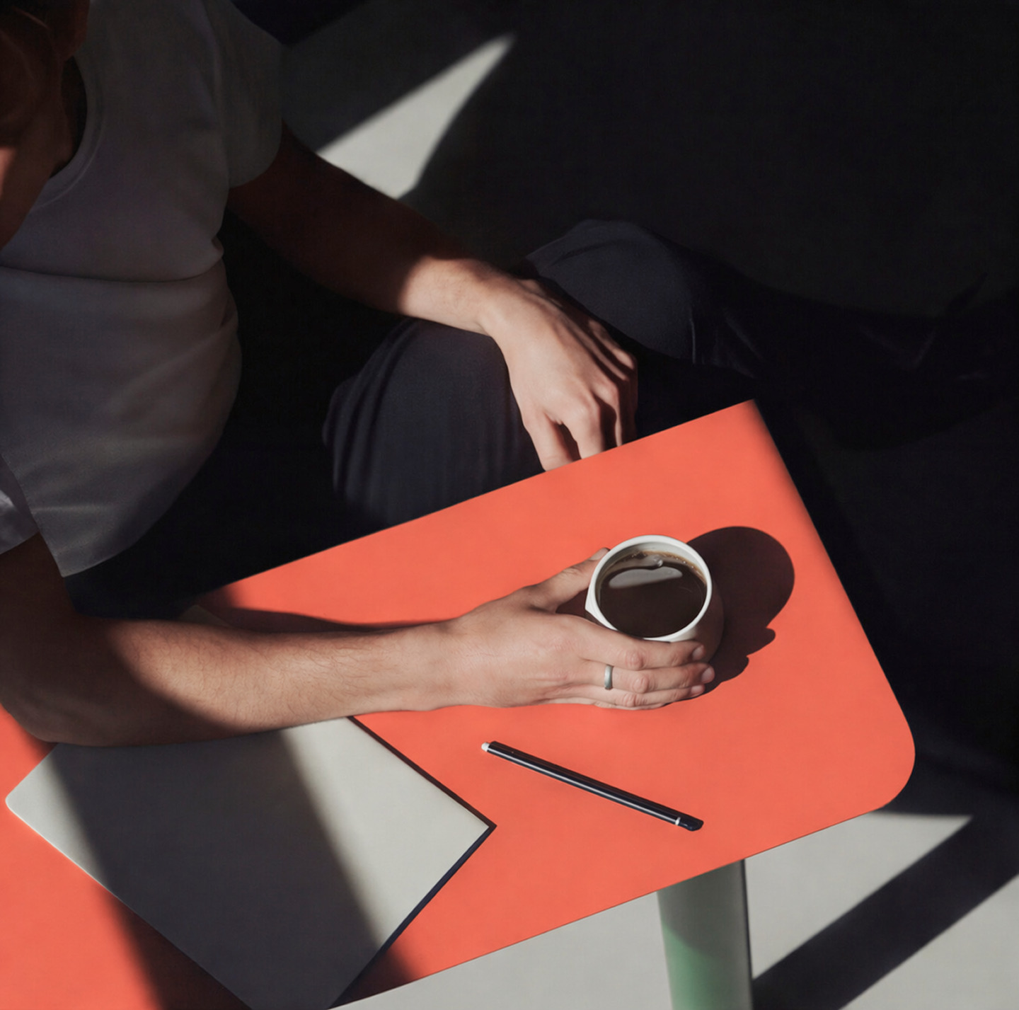 Close-up of a man's hands with a cup of coffee on an orange table next to a laptop and a pencil