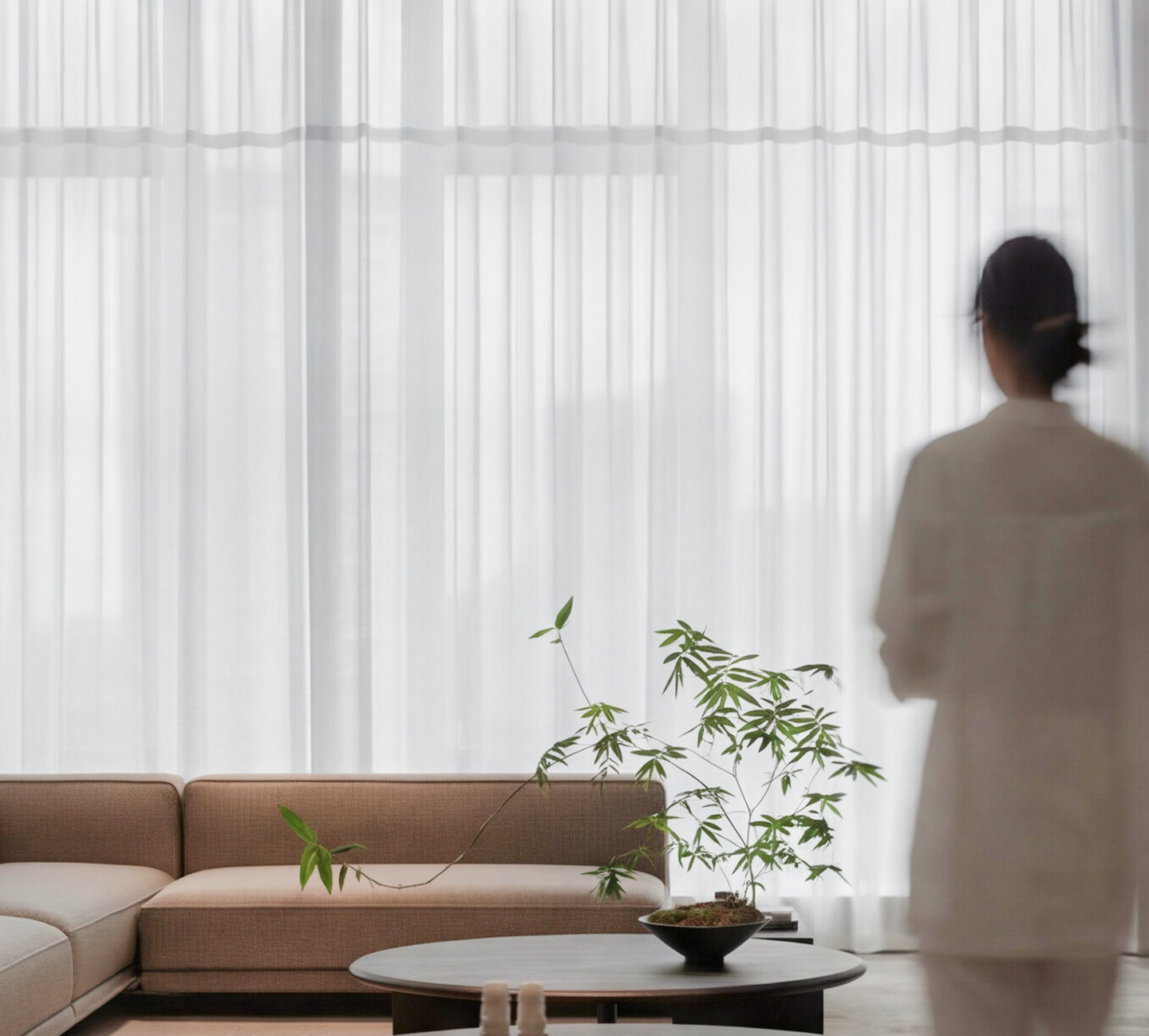 Minimalist interior with a beige sofa, a blurred figure by sheer curtains and a plant on a side table