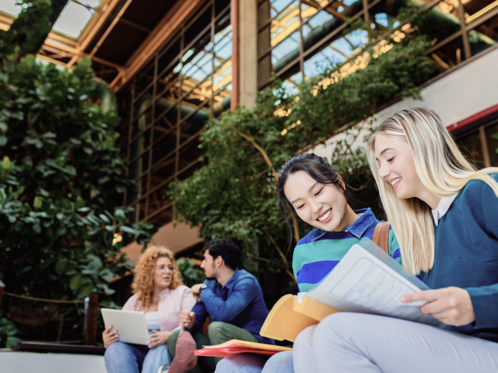 Two smiling female students looking through documents in a light-filled atrium with greenery