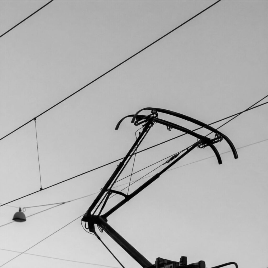 Black and white close-up of a tram pantograph and overhead lines against the sky