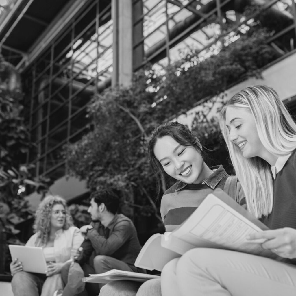 Smiling female students reading documents in a bright atrium with greenery