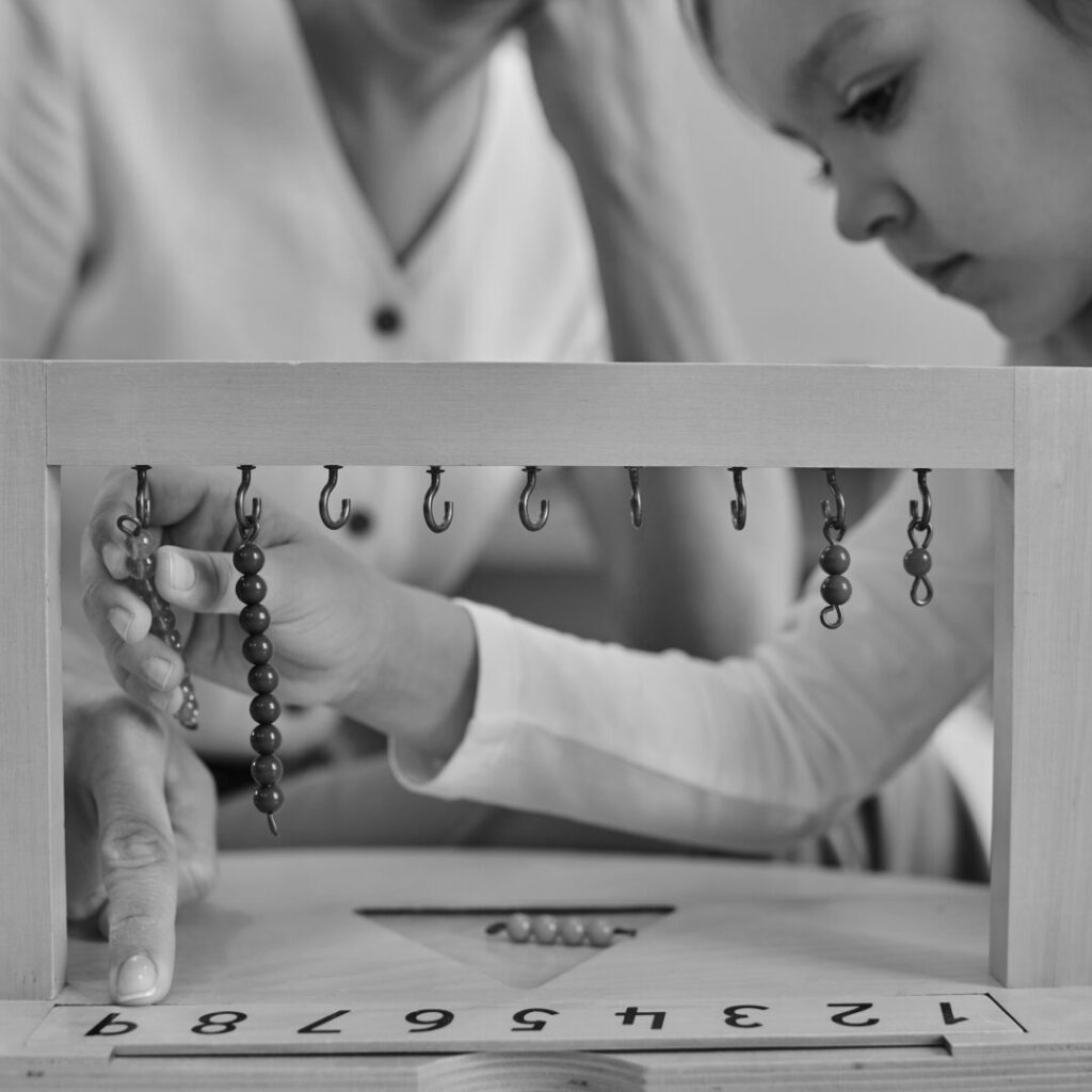 Black and white photograph of a child and an adult with a Montessori aid with beads and numerals