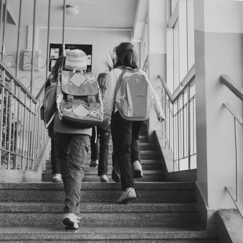 Black and white photograph of children with school backpacks climbing the stairs, seen from behind