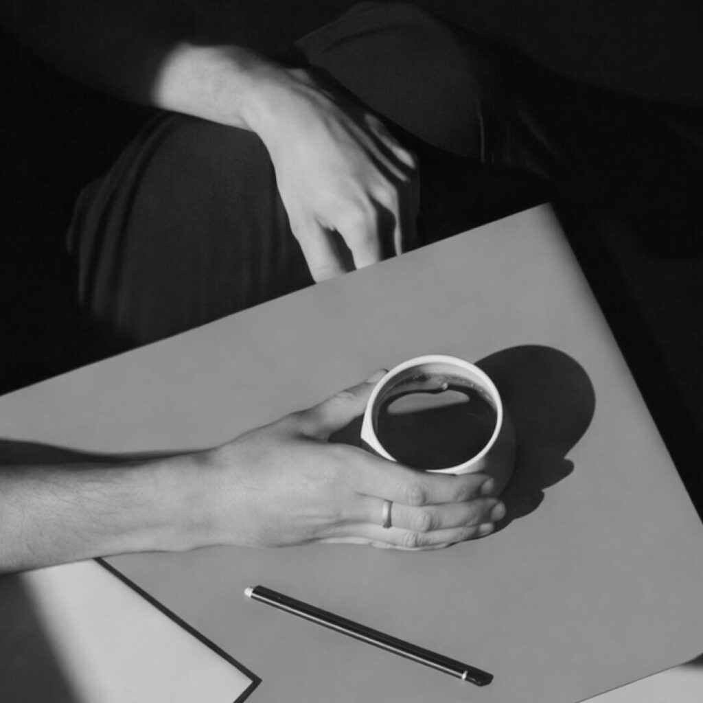 Black and white photograph of hands holding a cup of coffee at a table with a pen and paper