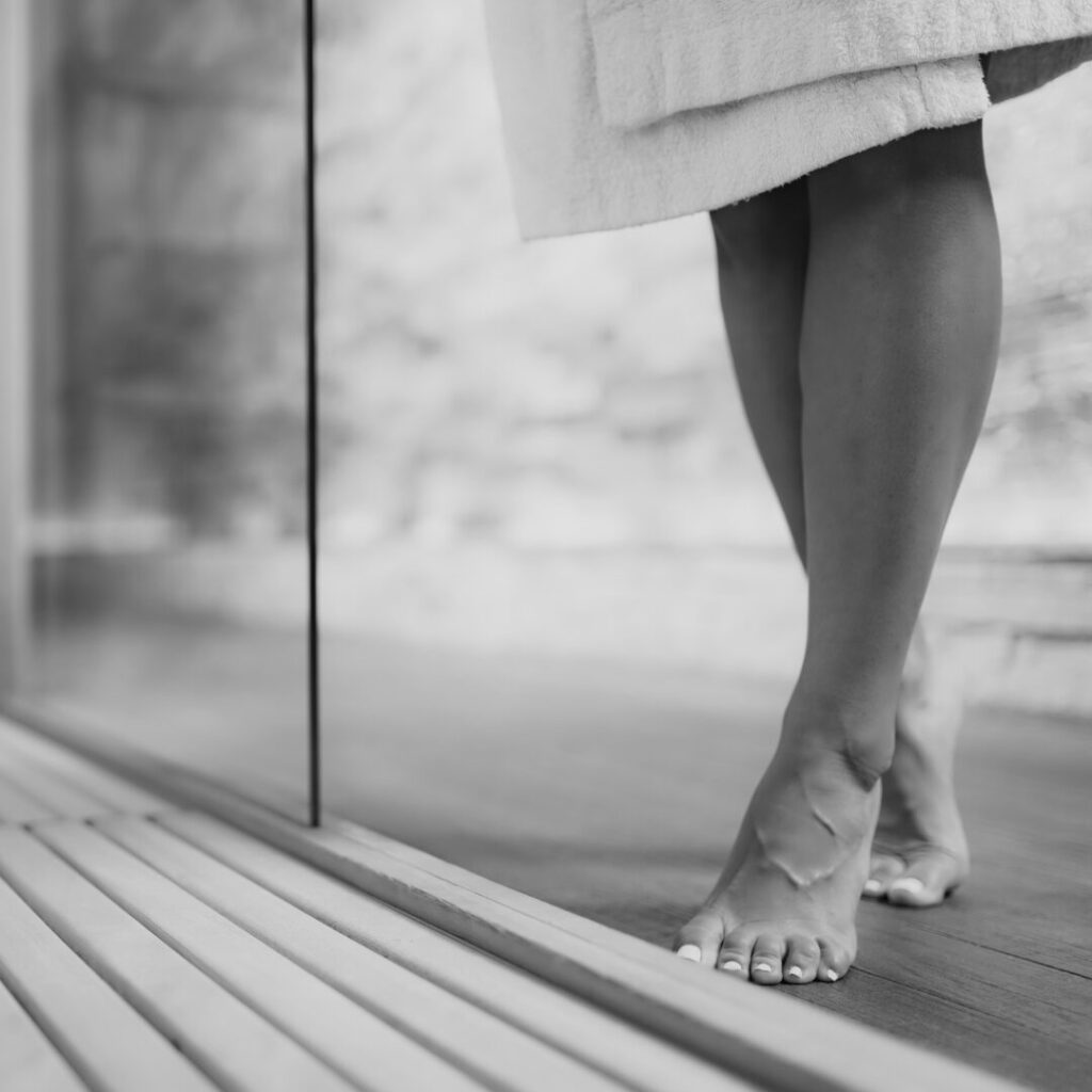 Black and white photograph of the bare feet of a woman in a robe on a wooden terrace by a glass wall