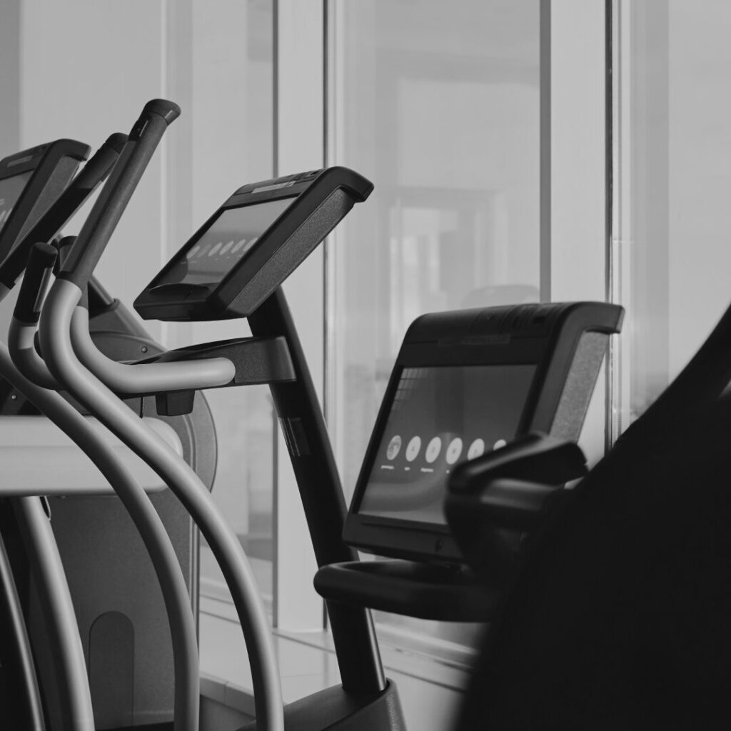 Black and white photograph of cardio machines with displays in a gym by a glass wall