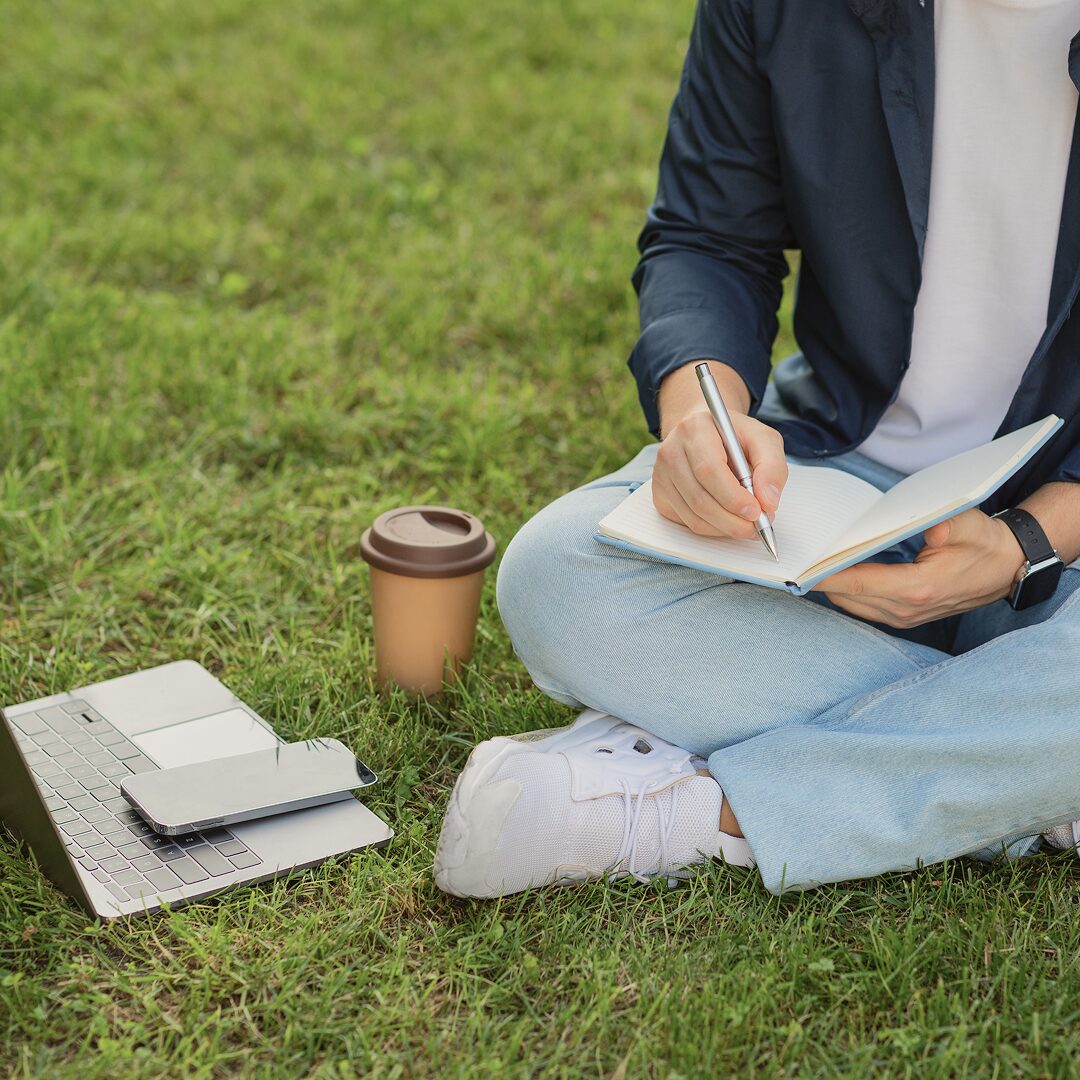 Young man sitting on the grass writing in a notebook next to a laptop and coffee